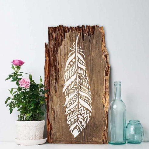 White feather stenciled onto a tall piece of bark edged reclaimed wood leaning by a rose and glass bottles