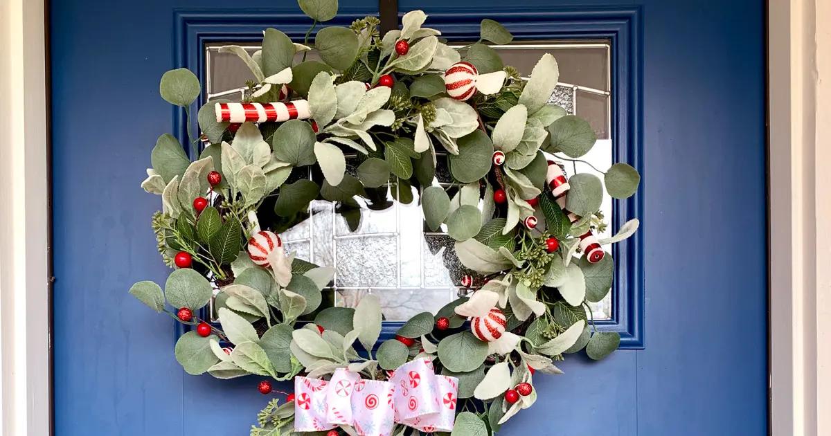 A eucalyptus wreath with peppermint candy ornaments, a striped candy stick, red berries and a pastel candy bow on a blue door