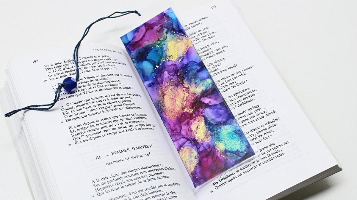 Alcohol ink bookmark in purple, blue and gold blooms with a navy beaded cord tassel resting on an open book