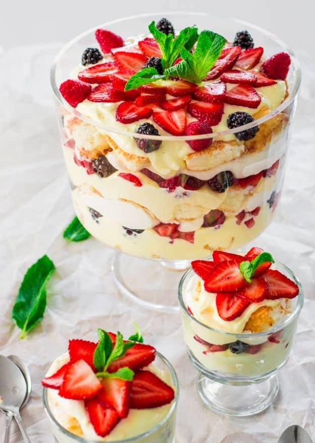 Glass trifle bowl layered with cake cubes, vanilla custard, strawberries and blackberries with small servings beside