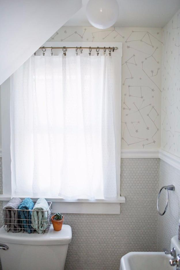 Sheer white no sew curtain panel clipped to a rod in a small bathroom window above a toilet and a towel basket