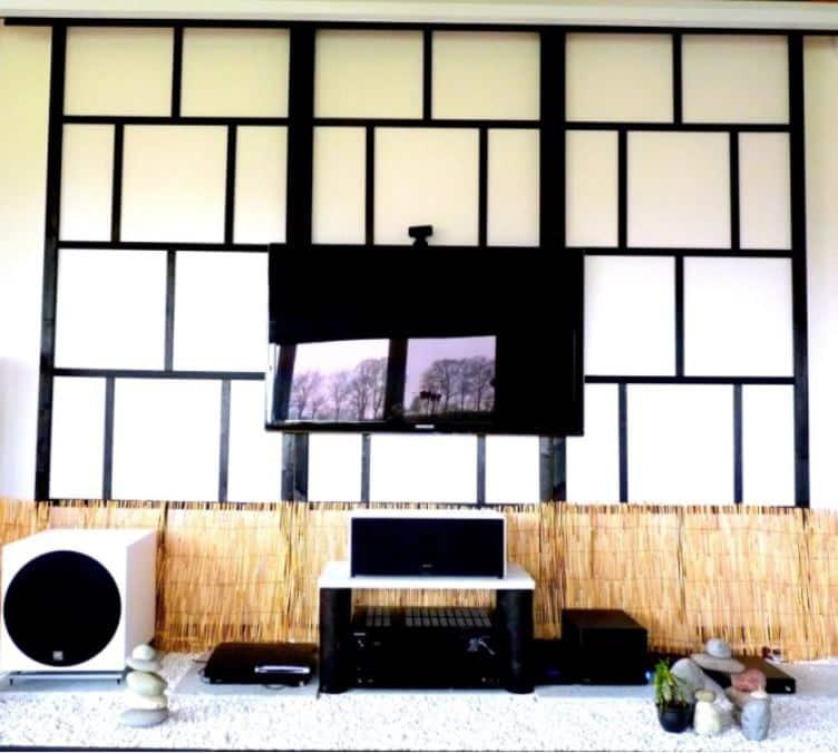 Wall of black framed shoji paper panels behind a wall mounted television, with a reed screen and pale gravel below