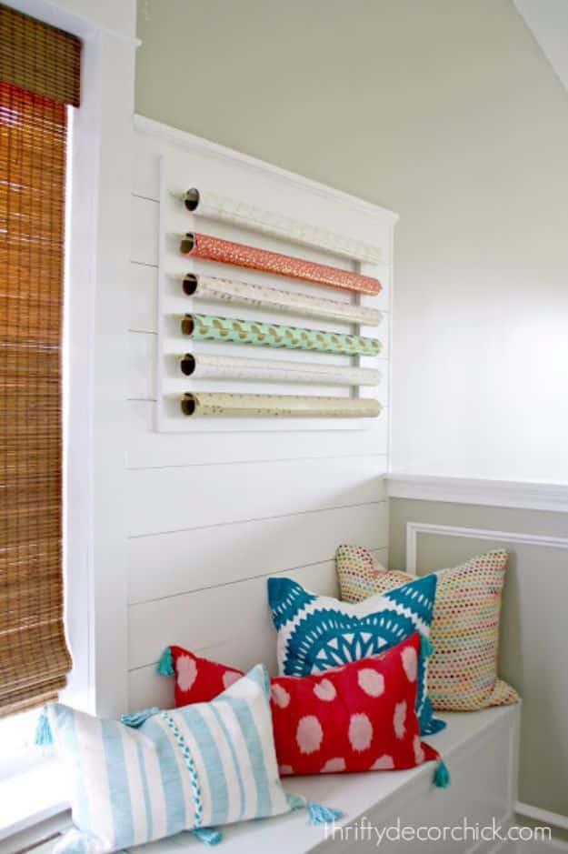 White wall frame above a window seat holding six rolls of wrapping paper on dowels, cushions and bamboo blind nearby