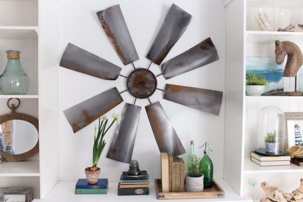 A large wooden windmill blade in weathered gray hangs on a white wall above a console table and a trailing plant