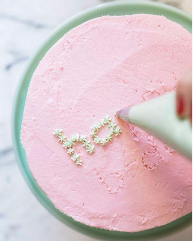 Pink frosted cake on a green stand with a piping bag adding green dots over toothpick marks spelling a word