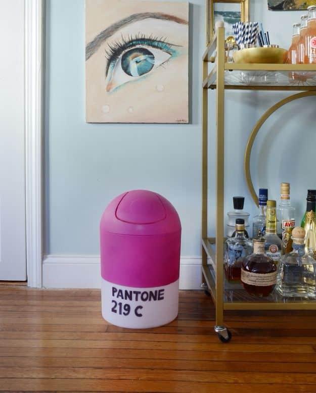Magenta and white swing lid trash can hand lettered PANTONE 219 C standing beside a gold bar cart on a wood floor