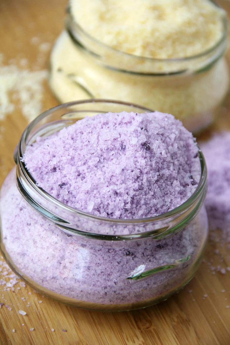 Two small glass jars of homemade bath salts, one lavender purple and one pale yellow, heaped full on a wood board