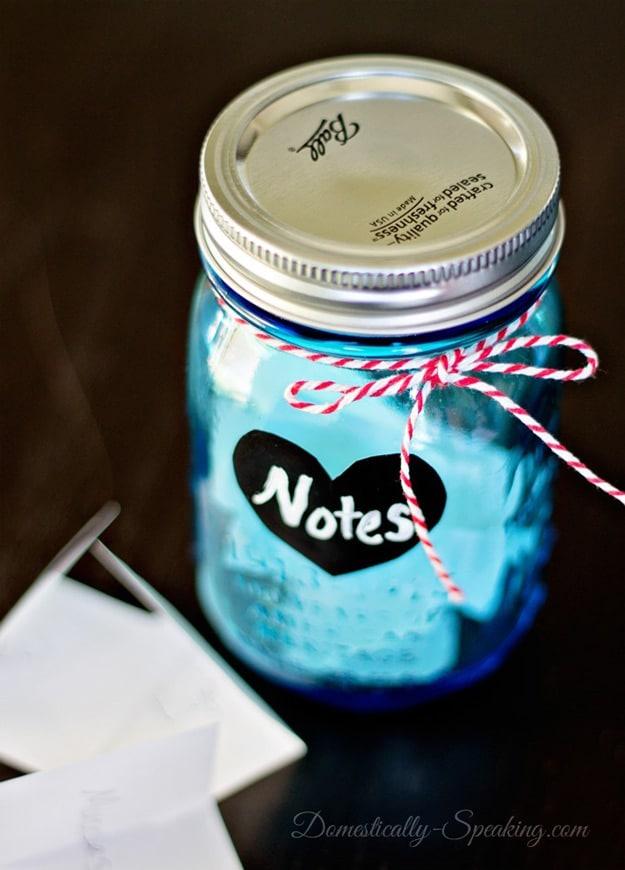 Wide mouth mason jar holding folded love notes with blank paper and a pen sitting beside it, an easy DIY gift to make at home