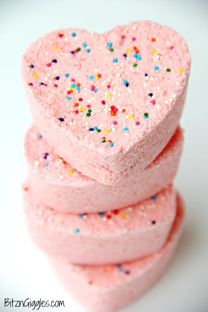 Stack of pink heart shaped bath bombs topped with rainbow nonpareil sprinkles on a plain white surface