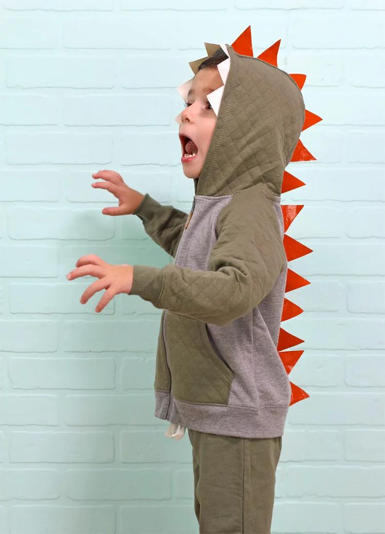 Child wearing a hooded orange dinosaur costume with white taped teeth and triangular spikes running down the back