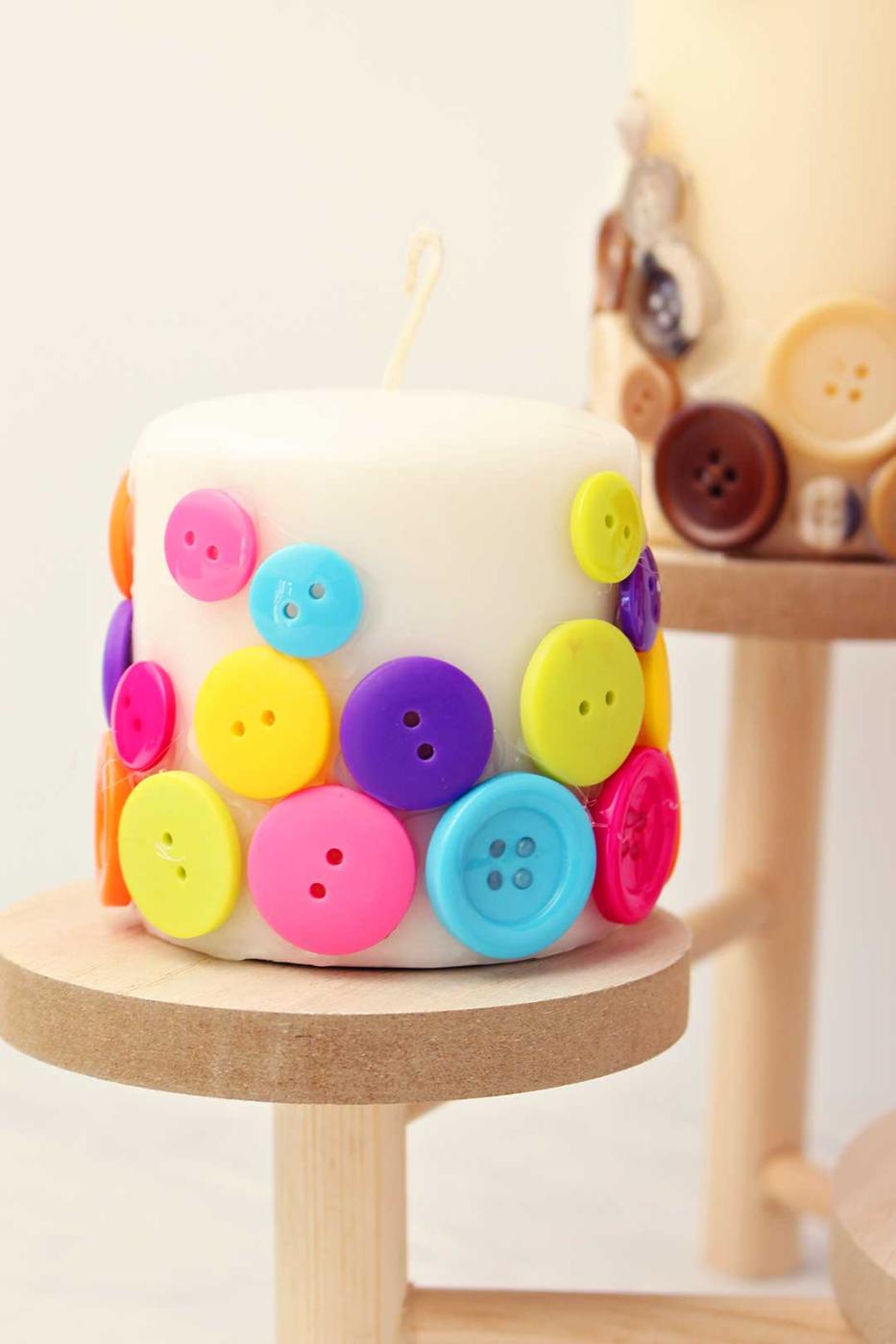 A white pillar candle covered in pink, blue, yellow and purple buttons on a small wooden stand beside a neutral button candle