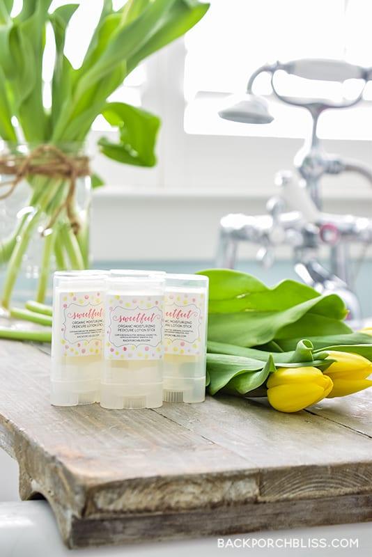 Three homemade lotion sticks in clear twist up tubes with printed labels on a weathered wood tray beside yellow tulips