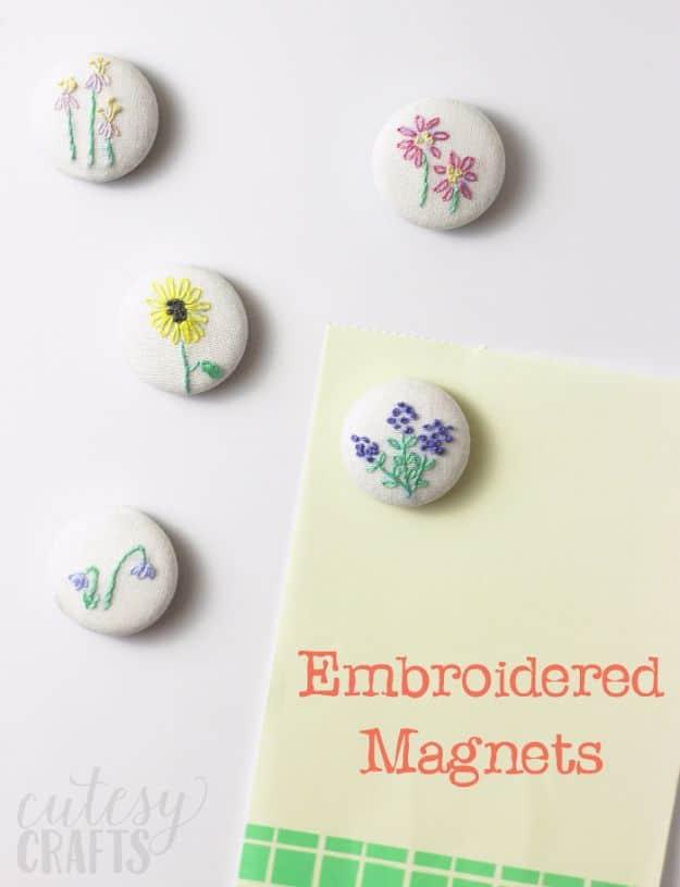 Five white fabric covered button magnets embroidered with small flowers, arranged on a white surface beside a card