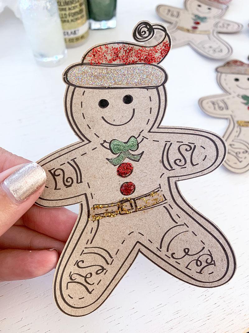 A hand holds a brown craft paper gingerbread man decorated with red, green and gold glitter glue and paint