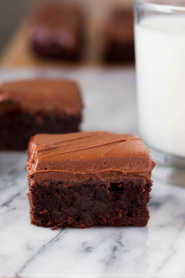 Fudge brownies with thick chocolate frosting on marble beside a glass of milk showing a dark dense crumb