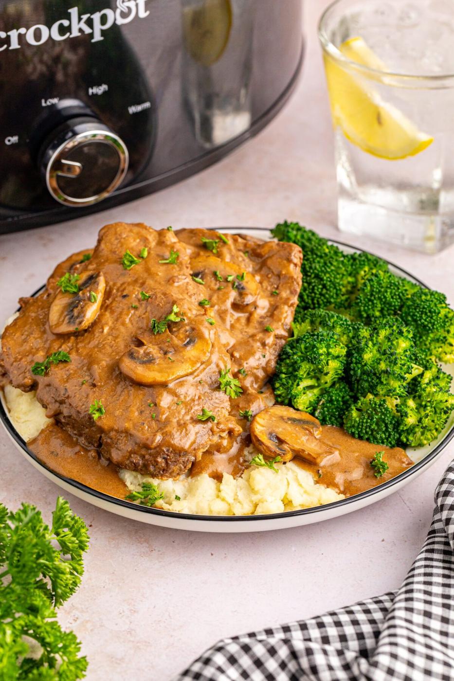 Cube steak in mushroom gravy over mashed potato in a dark rimmed bowl with broccoli and a black slow cooker behind