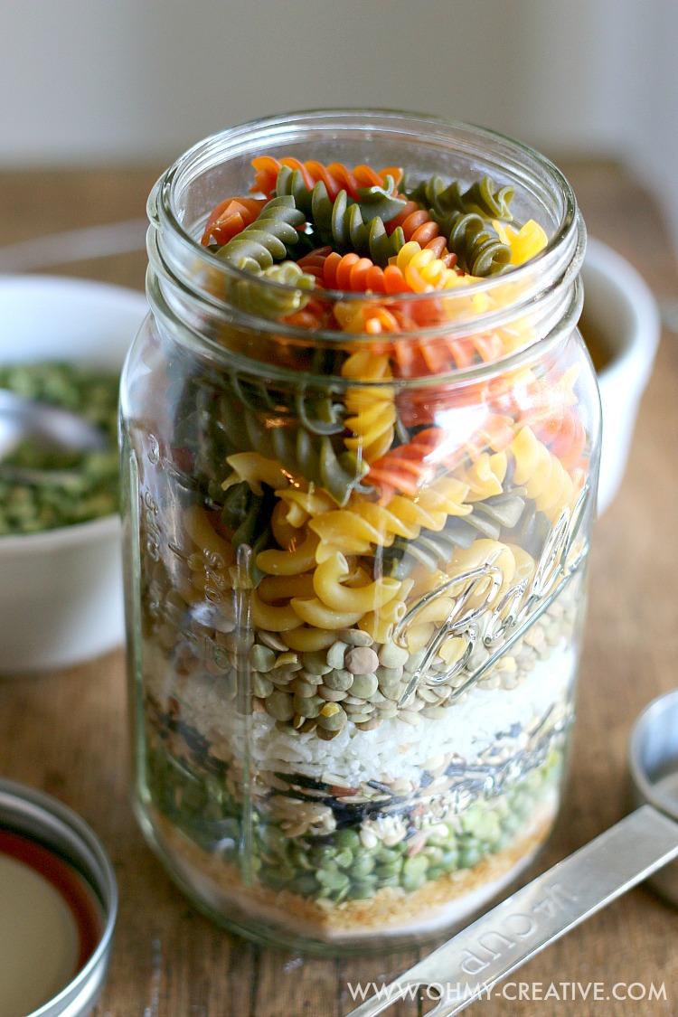 Wide mouth mason jar layered with tricolor rotini, lentils, split peas and rice on a wood table beside a lid