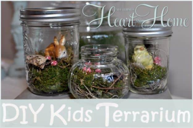DIY kids terrarium, four glass jars of green moss with tiny animal figures and pink flowers on a white windowsill