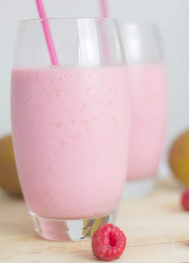 Two glasses of pale pink raspberry and pear smoothie with a pink straw beside a whole pear and fresh raspberries