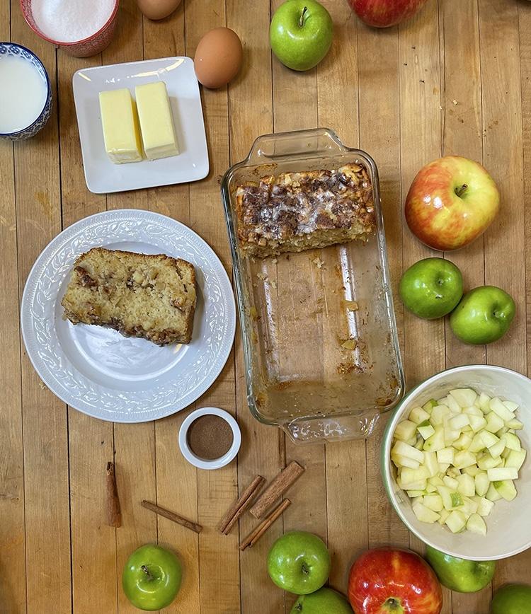 Homemade Apple Bread - glass loaf dish with several slices removed beside a plated slice, surrounded by butter, whole and diced apples, cinnamon sticks and milk