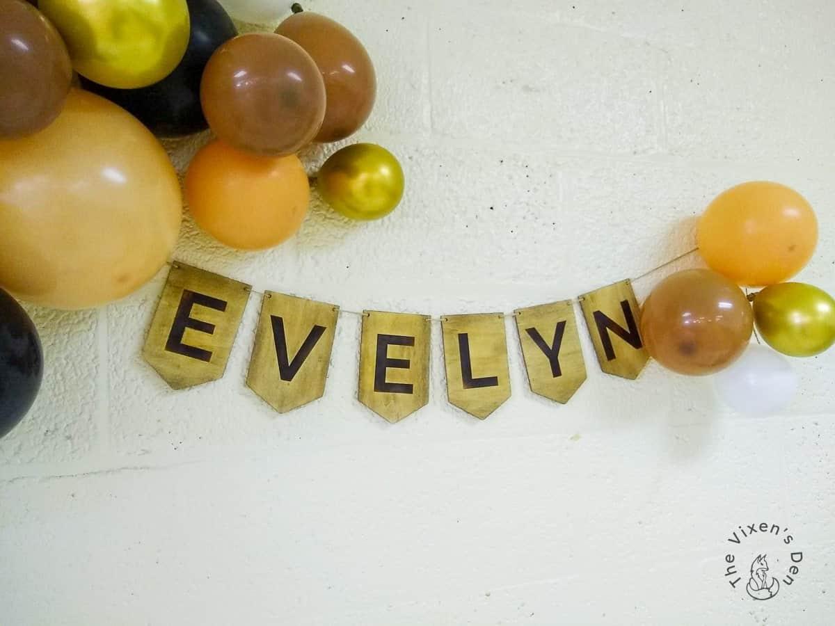 A wood pennant banner spelling Evelyn in burned letters hanging on a white brick wall among gold, brown and orange balloons