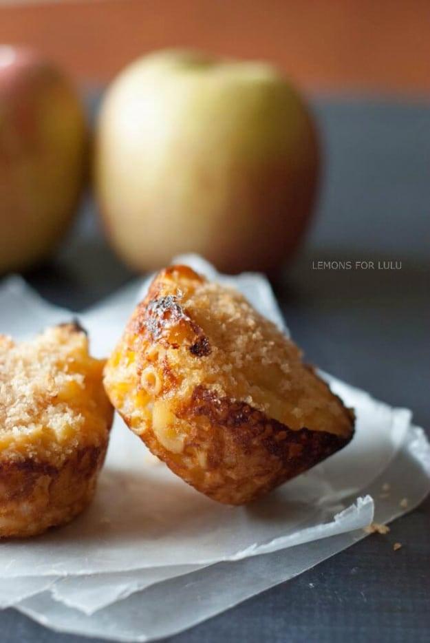 Two baked macaroni and cheese cups with crisp golden tops sitting on parchment paper beside two apples