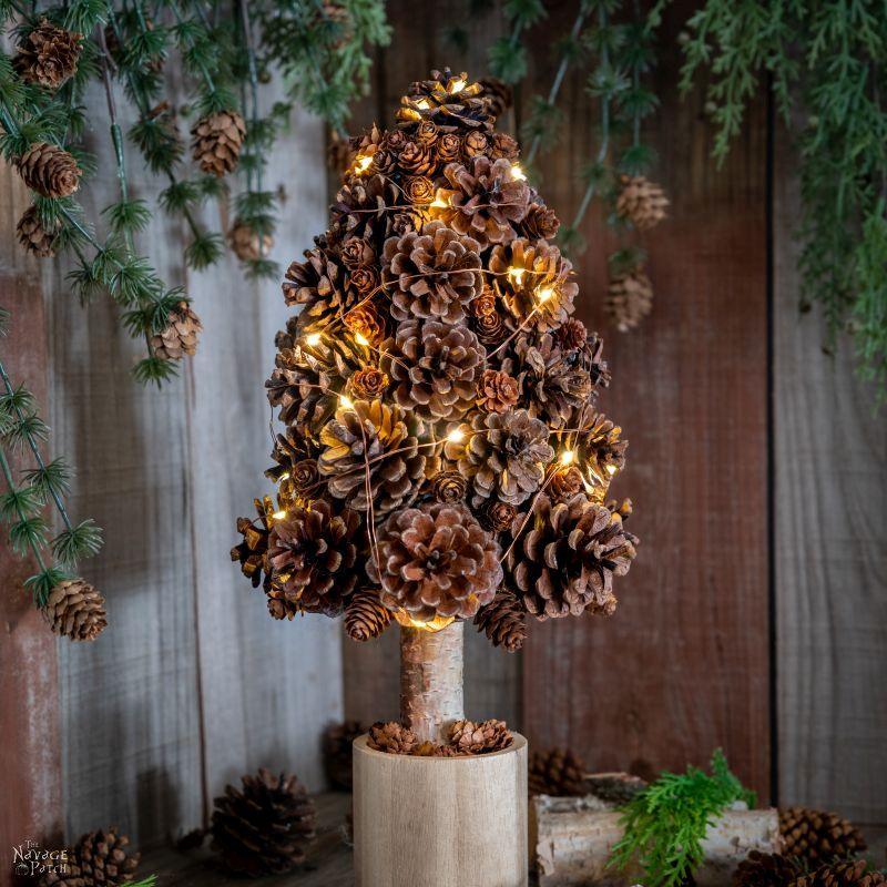 A tabletop tree made of pine cones on a birch trunk in a wooden pot, wound with glowing fairy lights against a wood wall