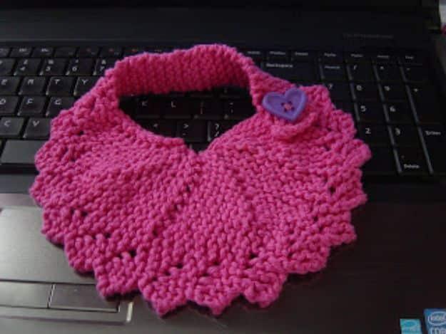 Bright pink knitted baby bib with a scalloped edge and small heart button, lying across a black laptop keyboard