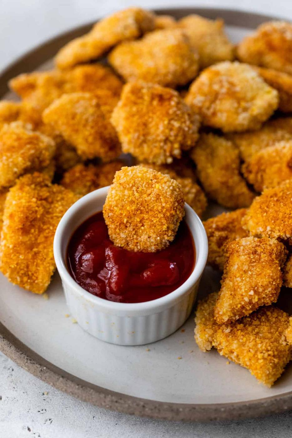 Baked chicken nuggets piled on a grey plate with a small white ramekin of ketchup beside them for dipping