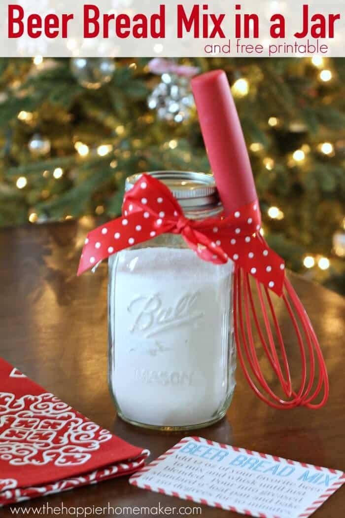 A quart mason jar of beer bread mix tied with red polka dot ribbon and a red whisk in front of a lit Christmas tree