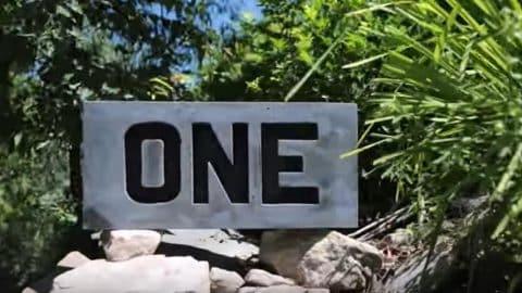 Concrete house number slab with the word ONE cast in raised black letters, set on rocks beside a path