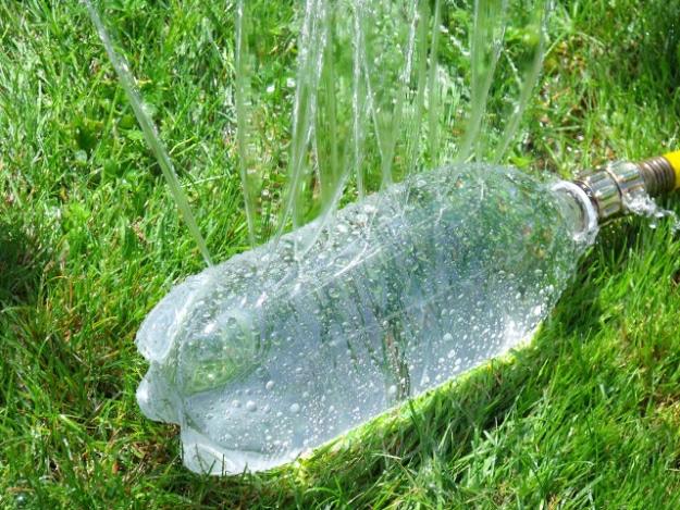 Clear two liter bottle pierced with holes lying in green grass, water spraying out in jets with a garden hose attached