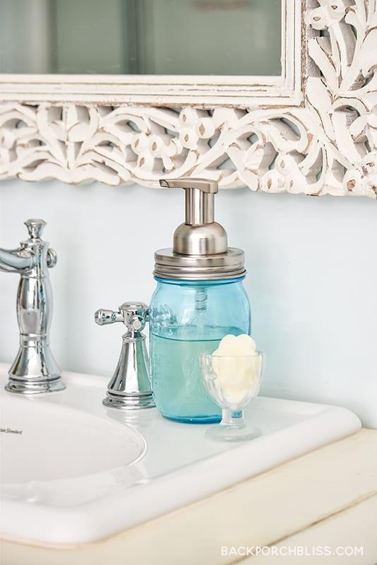 Blue pint mason jar of foaming hand soap with a brushed nickel pump on a white sink beside an ornate mirror