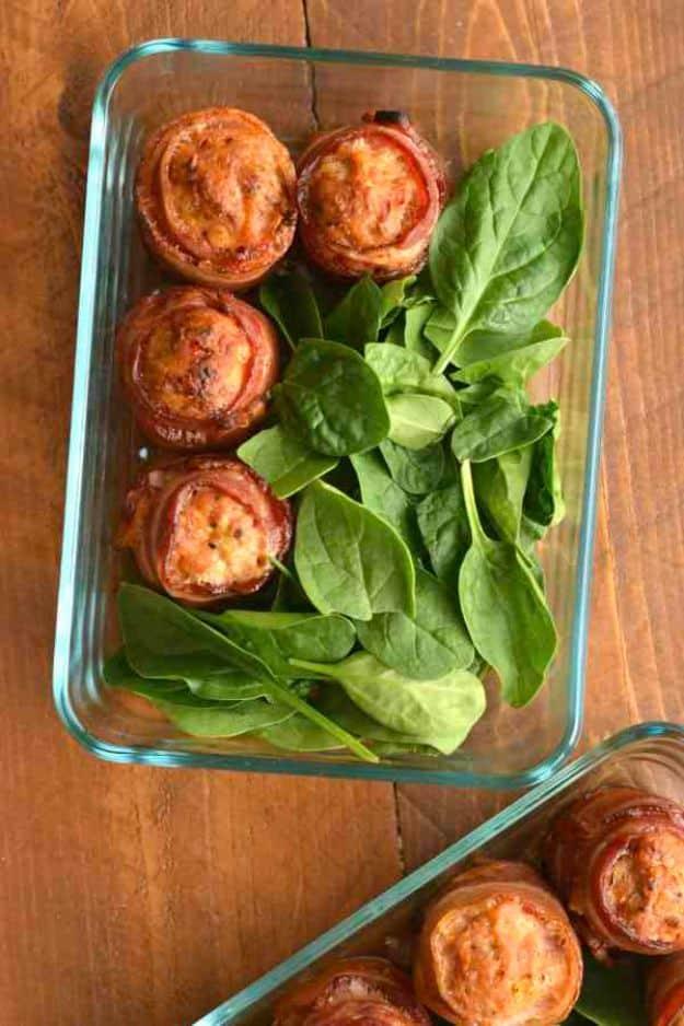 Keto bacon wrapped sausage meatballs in a clear glass dish with fresh spinach leaves alongside them on wood