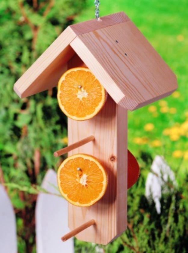 Cedar wood oriole feeder with peaked roof and dowel perches, two orange halves speared on nails, garden behind