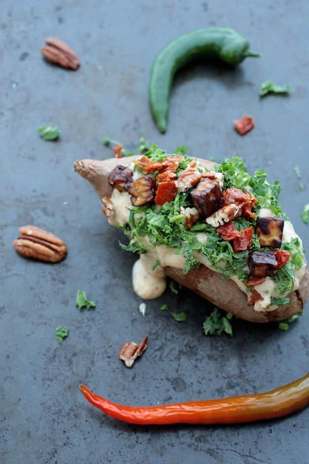 Stuffed sweet potato topped with cauliflower cheese sauce, kale, tempeh and pecans on a dark blue surface
