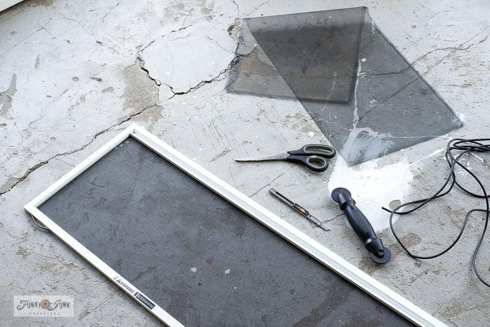 Old window screen frame laid on concrete with the torn mesh removed, scissors, spline cord and a spline roller beside it