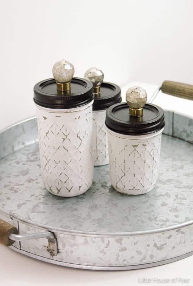 Three quilted mason jars painted white and distressed, with black lids and glass knobs, on a galvanized tray