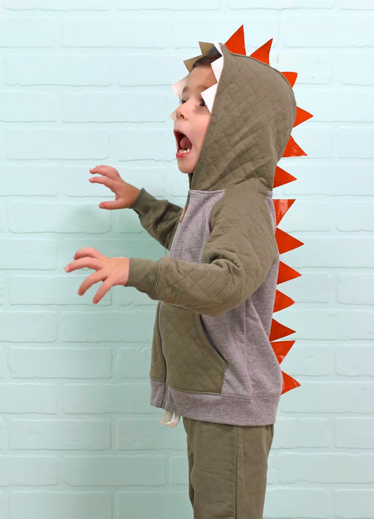 Child wearing a gray-green hooded dinosaur costume with white taped teeth and red-orange triangular spikes down the back