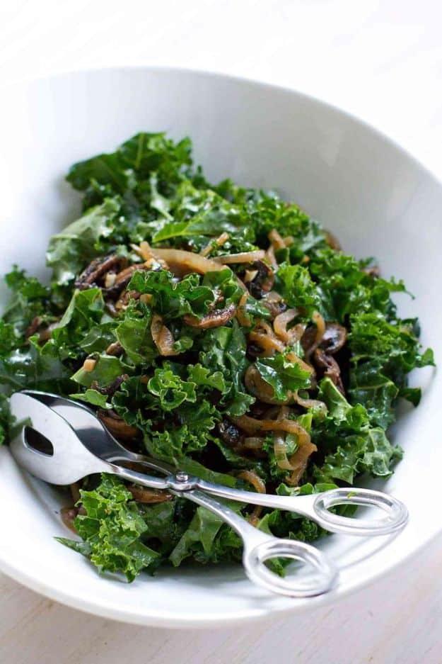 Warm kale salad with caramelized onions and sliced mushrooms tossed with serving spoons in a white bowl