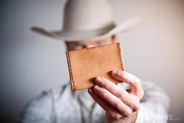 Simple leather wallet, a tan hand stitched leather billfold held up to the camera by a man in a pale cowboy hat