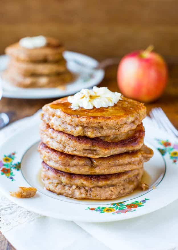 Apple pie pancakes stacked on a floral plate with whipped cream on top and a red apple in the background