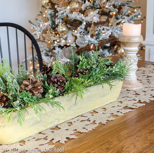 A long wooden dough bowl filled with fresh cedar, spruce and boxwood clippings and pinecones on a table beside a flocked tree