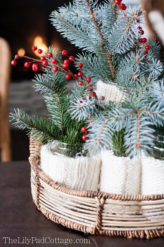 Glass jars wrapped in cream sweater sleeves holding blue spruce and red berry stems in a round wicker tray by a fireplace