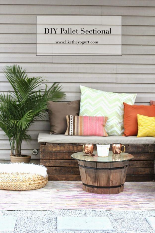 DIY pallet sectional with a long cushion and bright pillows against gray siding beside a barrel coffee table