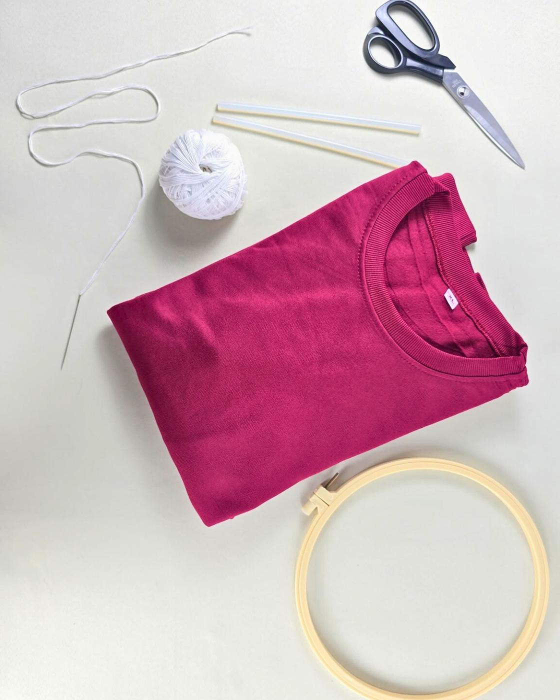 DIY Embroidery - flat lay of a folded burgundy knit sweater with white cord, a ball of white yarn, an embroidery needle, hot glue sticks, scissors, and a wooden hoop