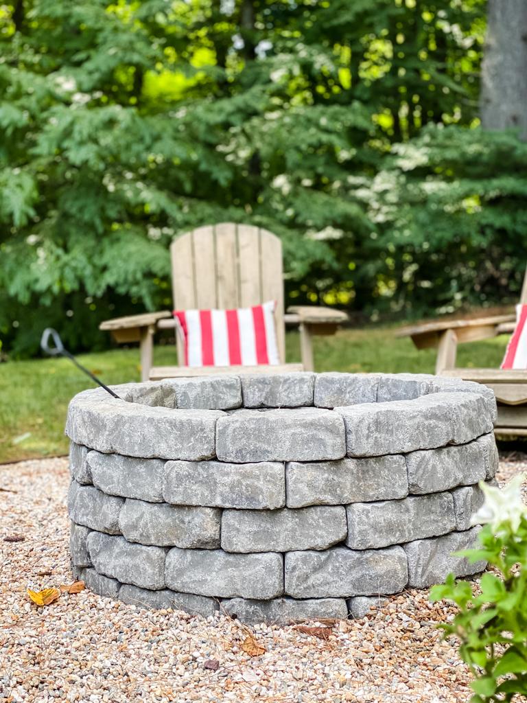 DIY paver fire pit, a stacked gray stone ring set on pea gravel with wooden Adirondack chairs and striped pillows