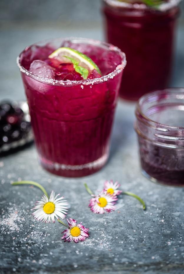 Deep purple blueberry basil margaritas in salt rimmed glasses with daisies and blackberries on slate