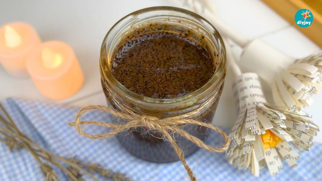 DIY coffee body scrub in a glass jar tied with twine, sitting on blue gingham with candles and a rolled paper fan