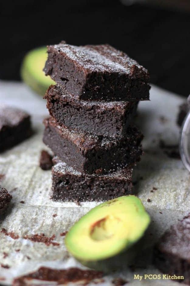 Three stacked dark avocado keto brownies on a pale white surface with a halved ripe avocado beside them
